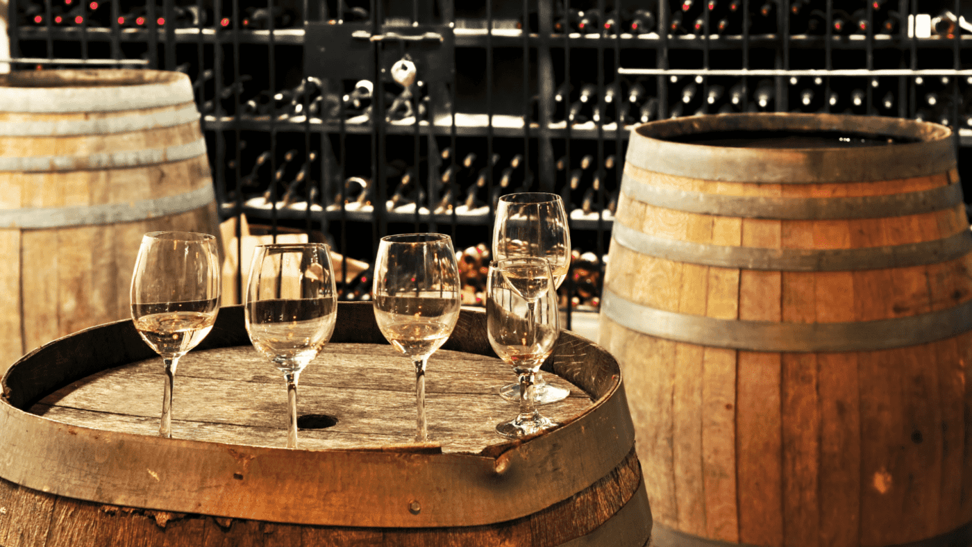 In a wine cellar, rows of wine rest on a stand, in front of it is a barrel with wine glasses.