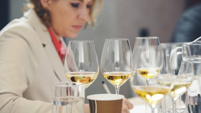 A woman in a lab coat sits in front of several glasses of wine and takes notes.