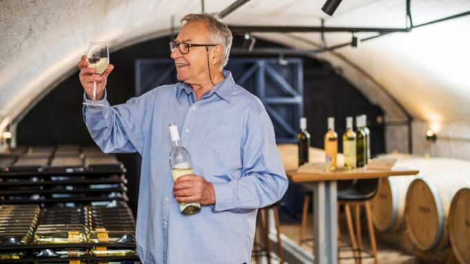A winemaker holds a bottle of white wine in one hand and a glass of wine in the other.