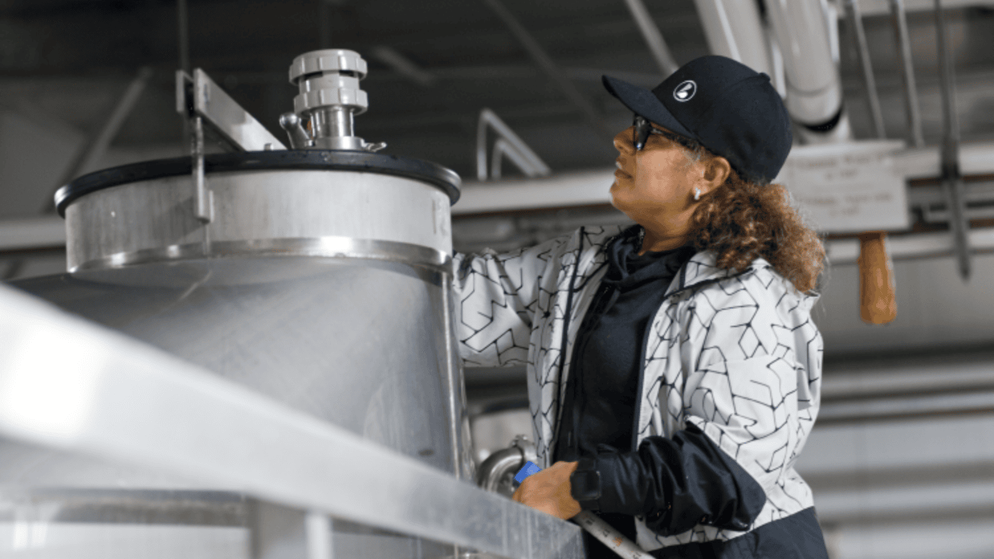 A BevZero technician stands on a ladder to reach the top of a wine tank for measurements.